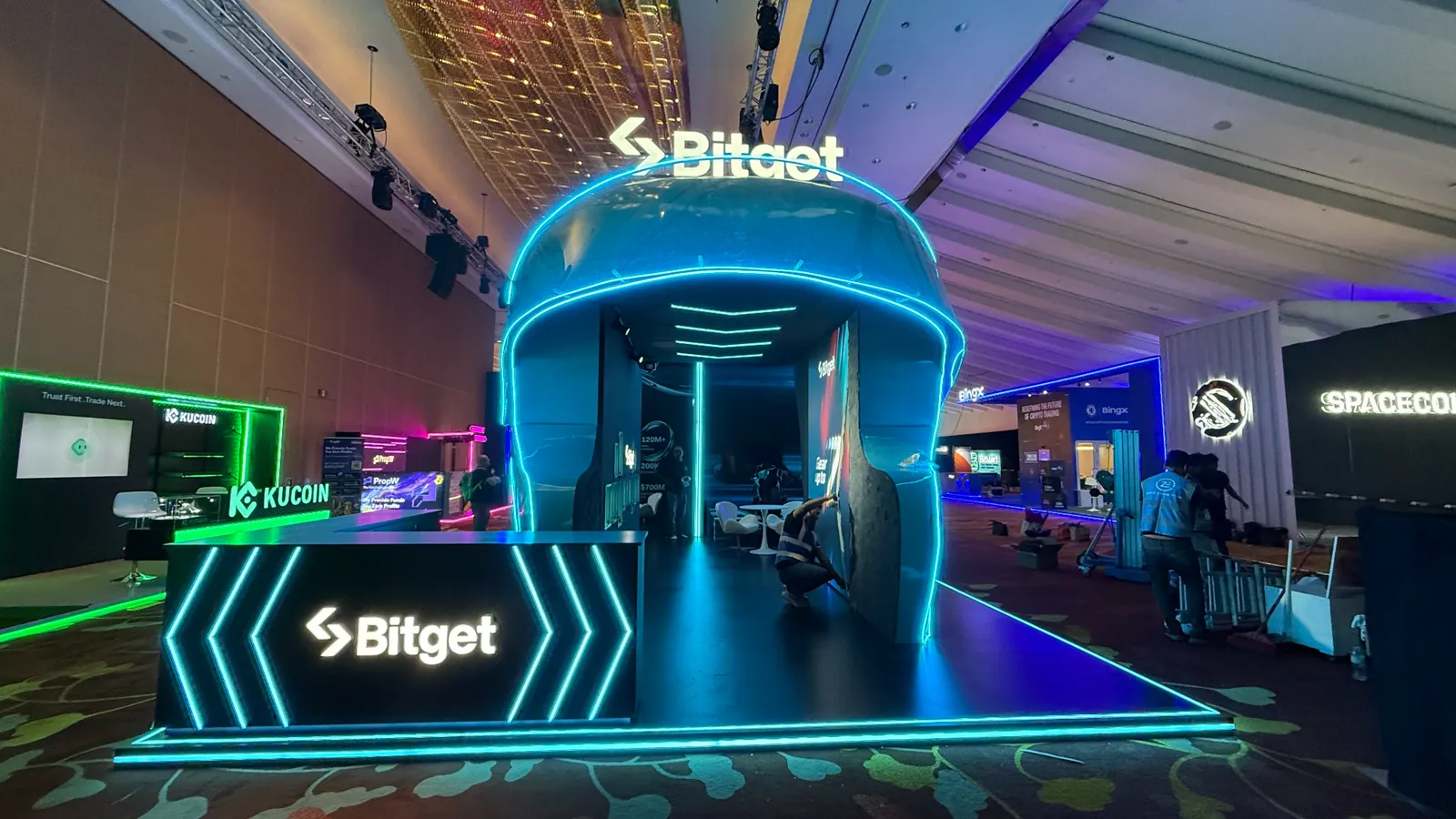 Completed Bitget exhibition booth at TOKEN2049 with a reflective blue curved shell and cyan edge lighting